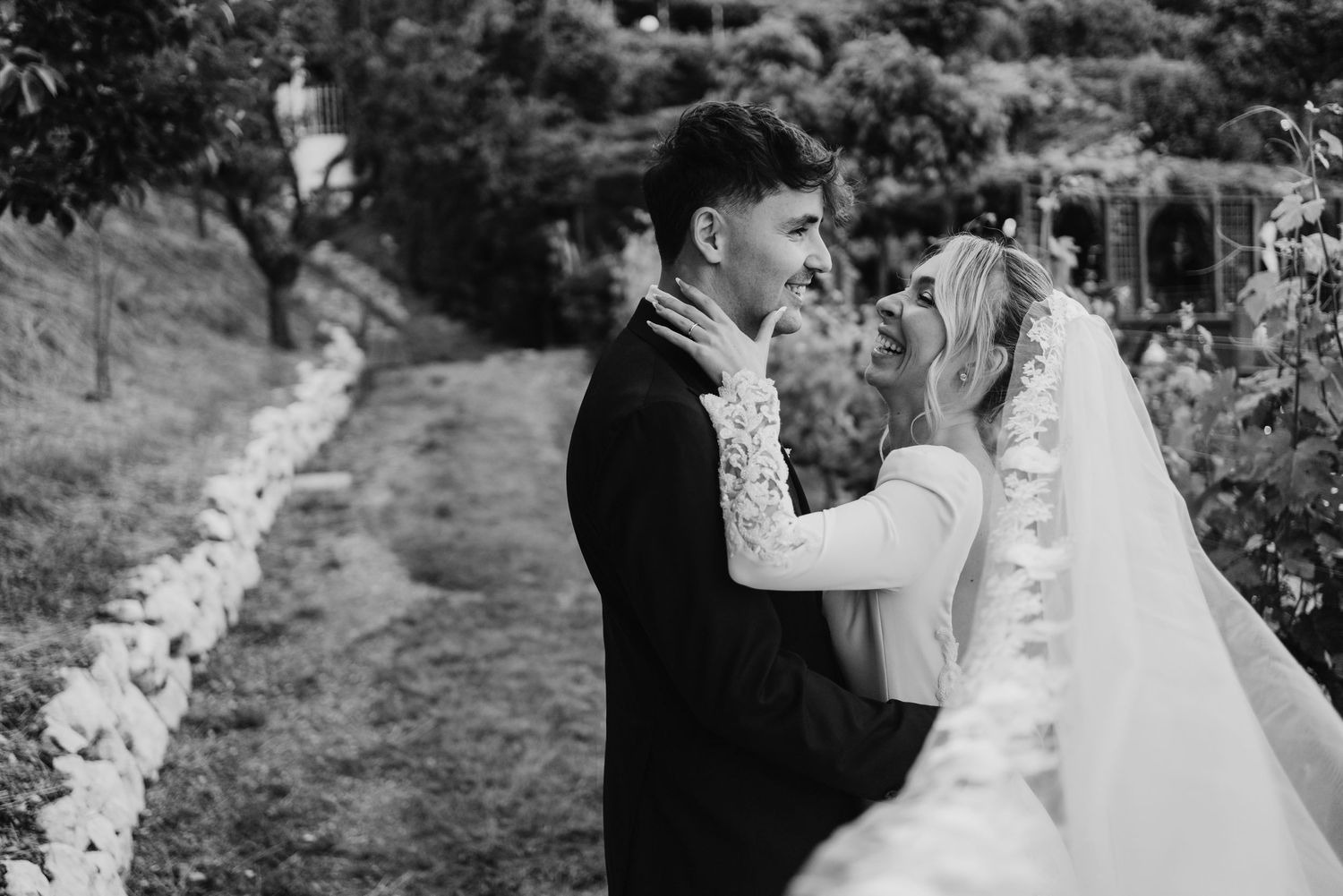 Black and white photo of smiling bride and groom embracing along stone-lined vineyard path.