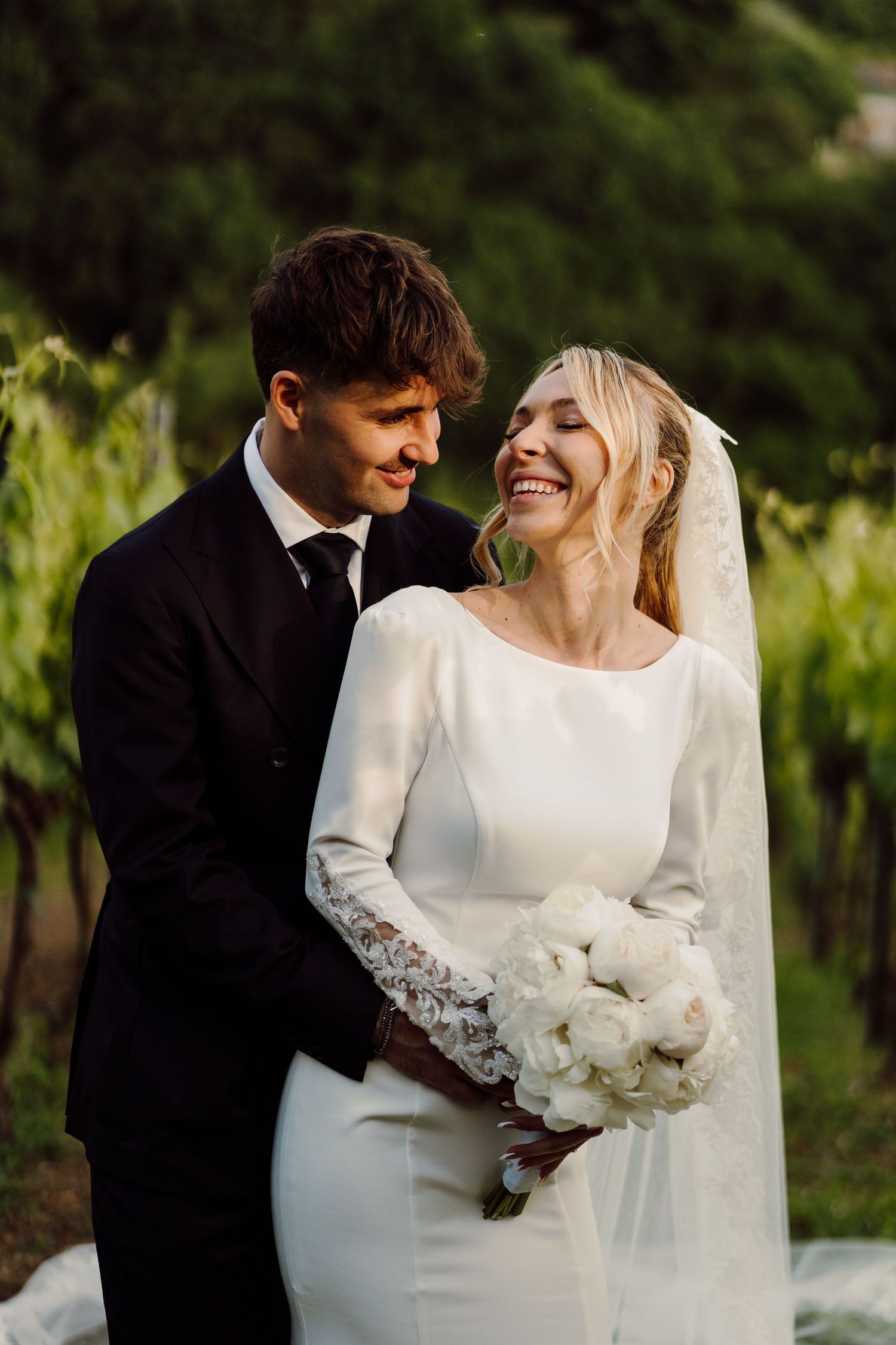 Joyful bride in minimalist white gown holding peony bouquet, laughing with groom in black suit in vineyard.