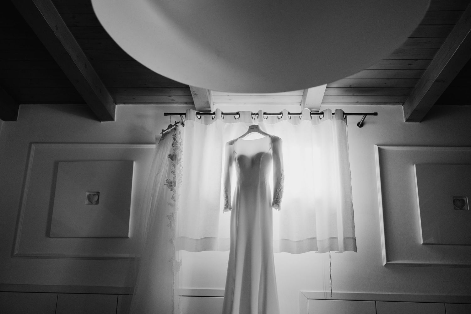 Elegant white wedding dress hanging on curtain rod in black and white photo.