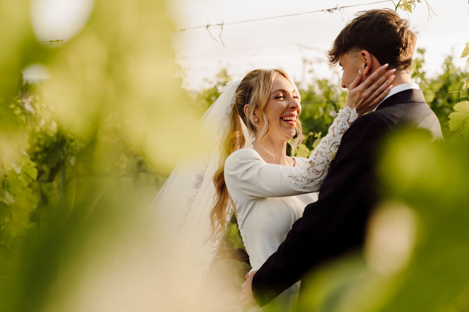 Radiant bride smiling at groom framed by lush green vineyard leaves in warm sunlight.