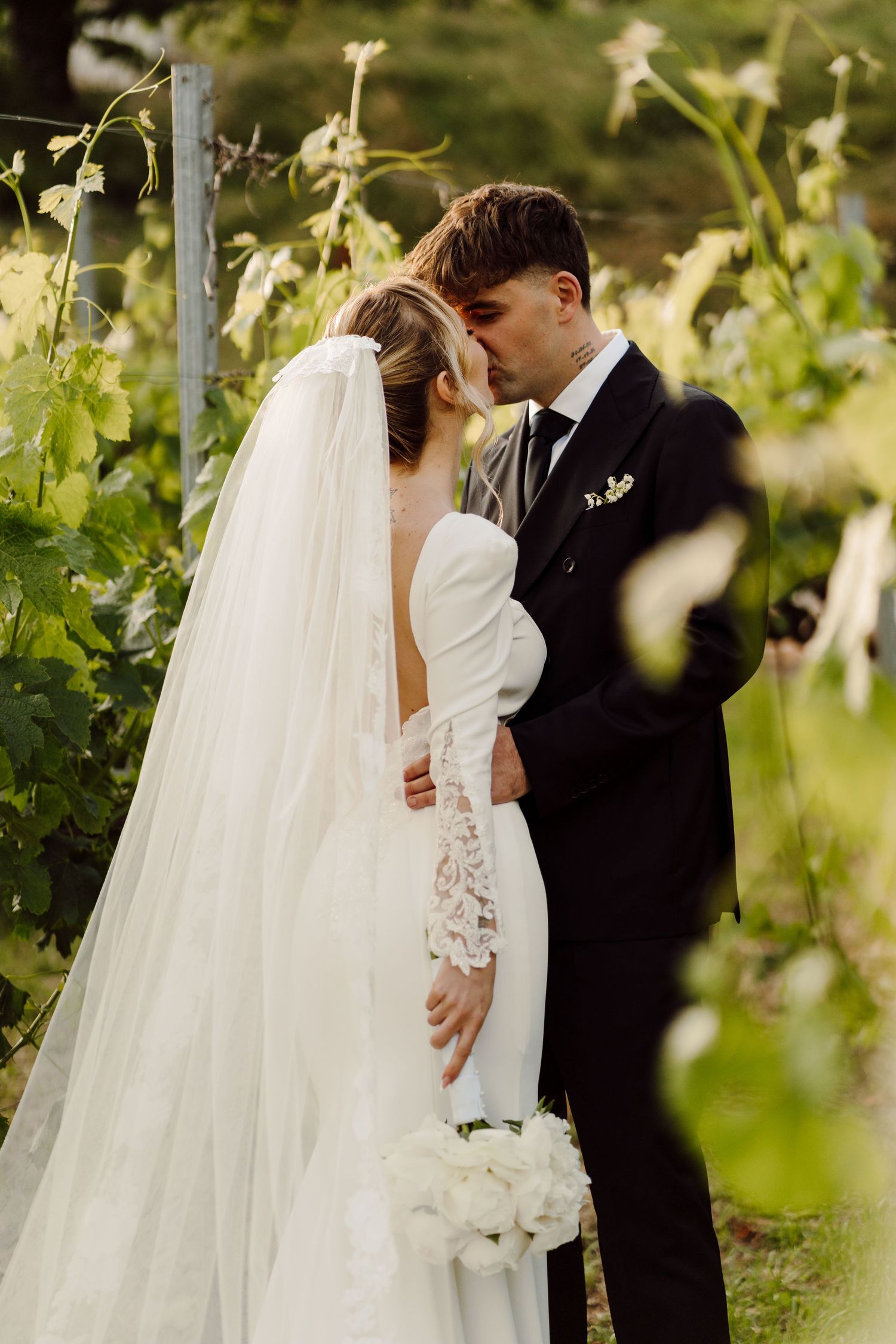 Groom kissing bride on forehead in sunlit vineyard, bride holding white peony bouquet in lace-sleeved gown.