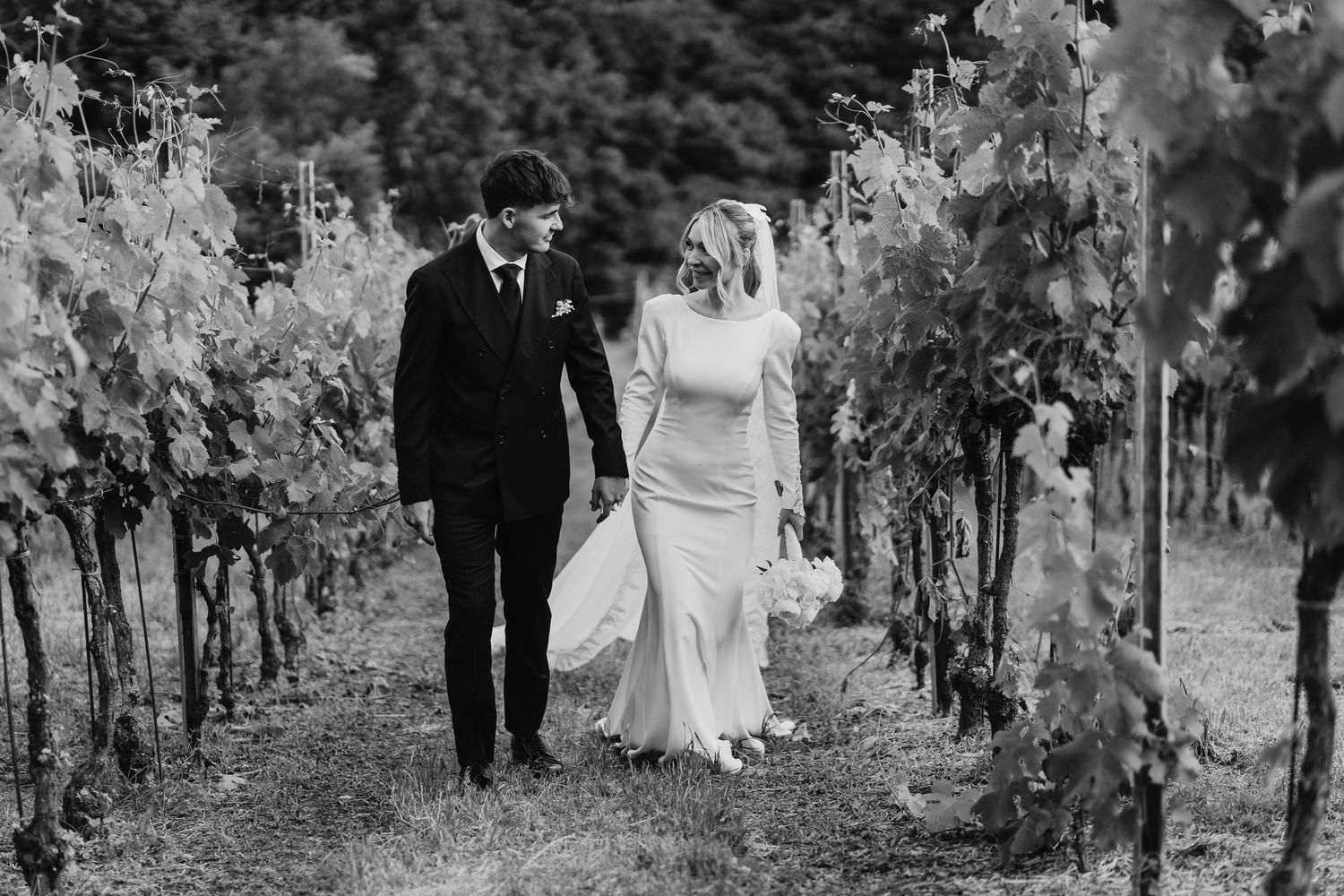 Black and white photo of bride and groom walking hand in hand through lush vineyard rows.