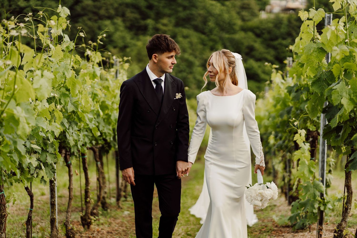 Bride and groom holding hands and gazing at each other while walking through a green vineyard.
