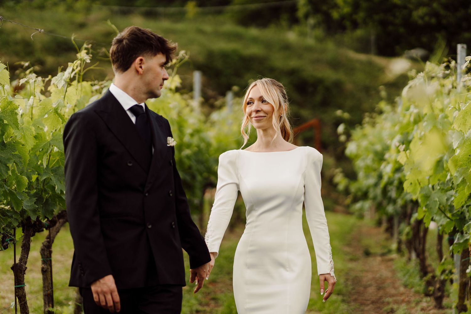 Groom in black suit and bride in white gown walking through vineyard rows on a cloudy day.