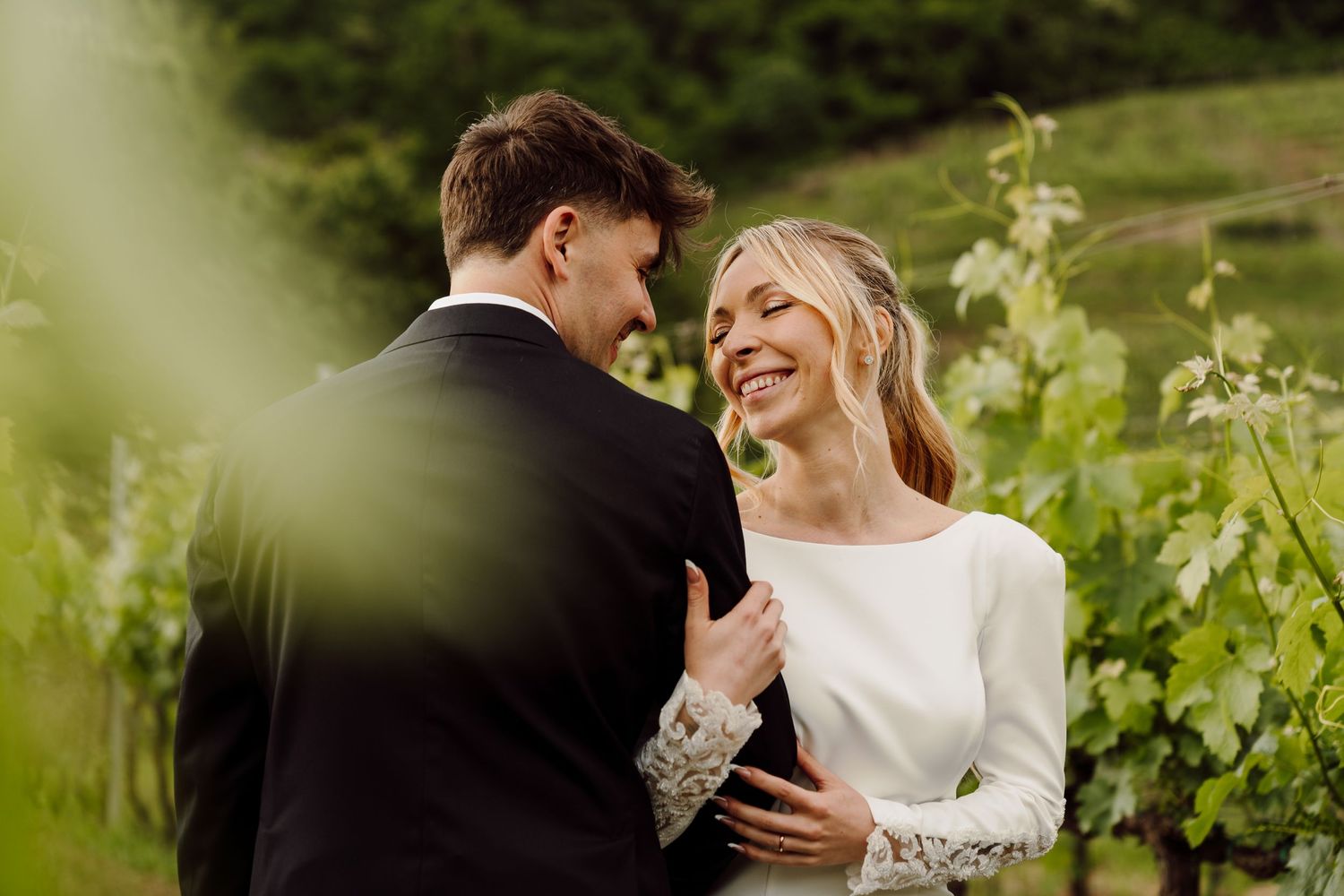Joyful bride laughing while embracing groom outdoors in a lush green vineyard setting.