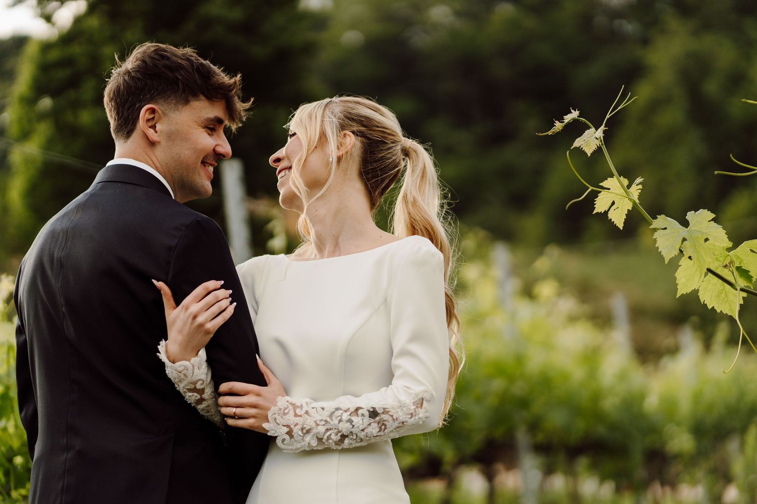 Smiling bride and groom embracing each other lovingly among green vineyard vines outdoors.