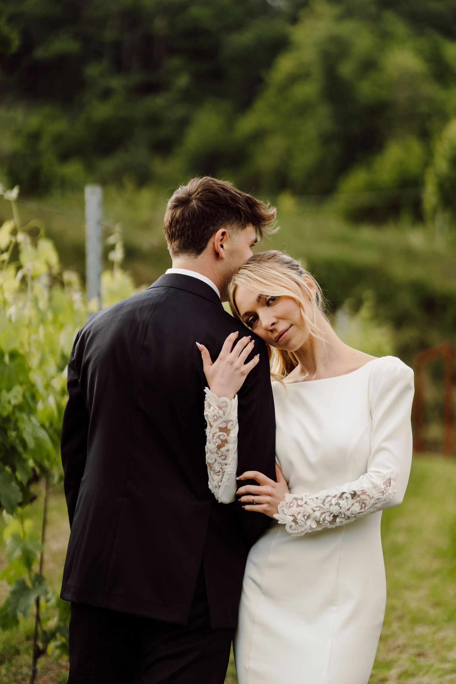 Bride in lace-sleeved gown leaning against groom in black suit in a scenic vineyard.