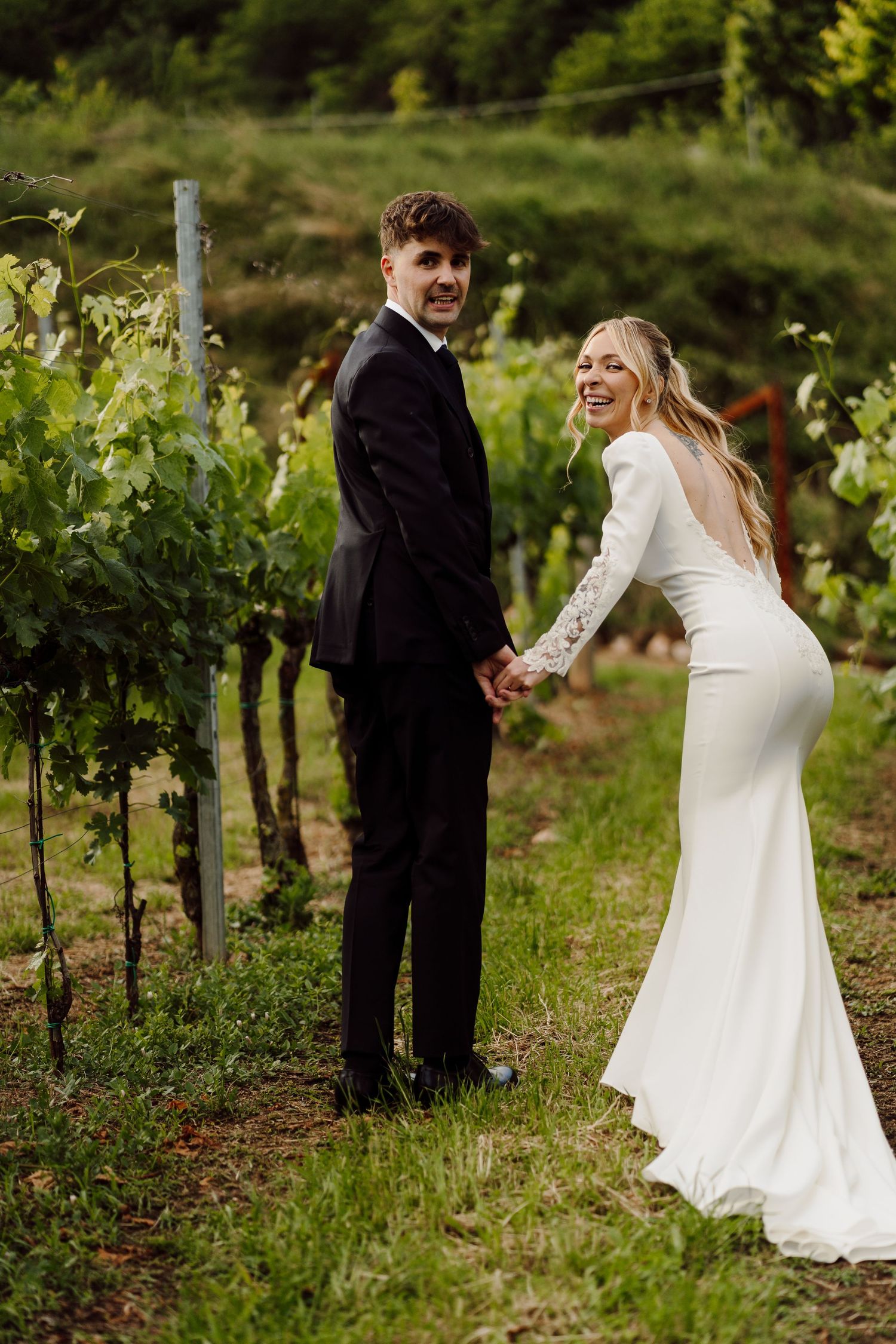 Bride and groom holding hands among vineyard rows, she in fitted white gown, he in black tuxedo.
