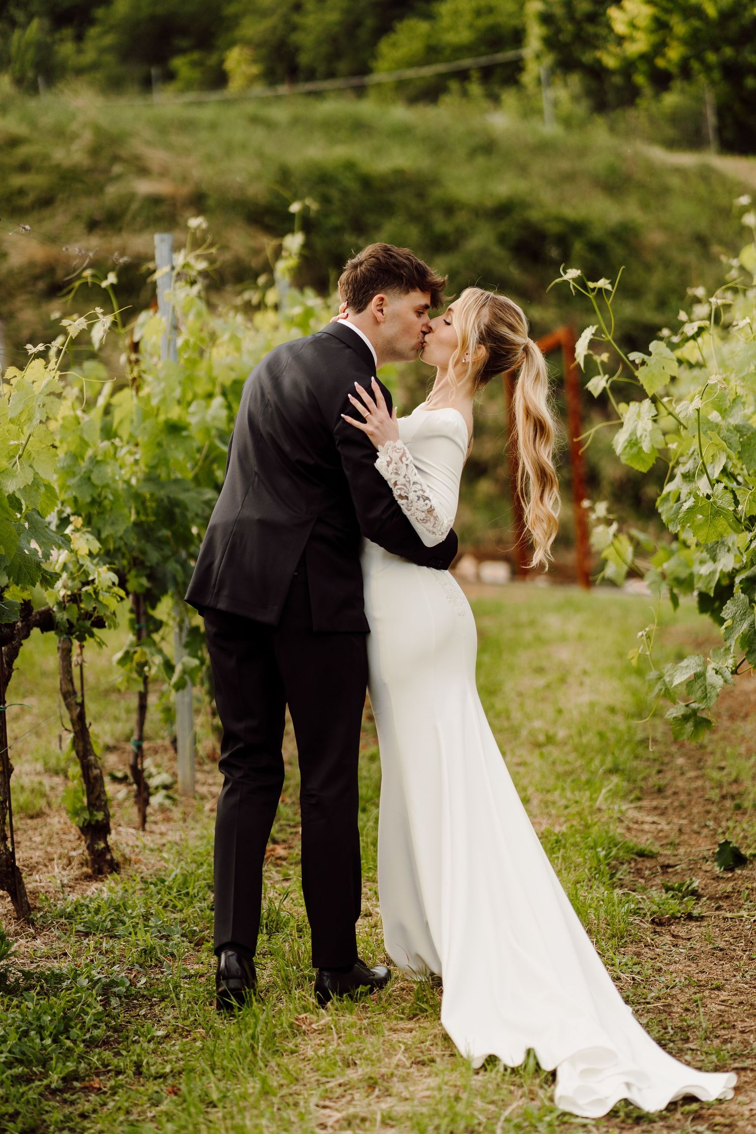 Newlyweds sharing a romantic kiss in a lush green vineyard on their wedding day.