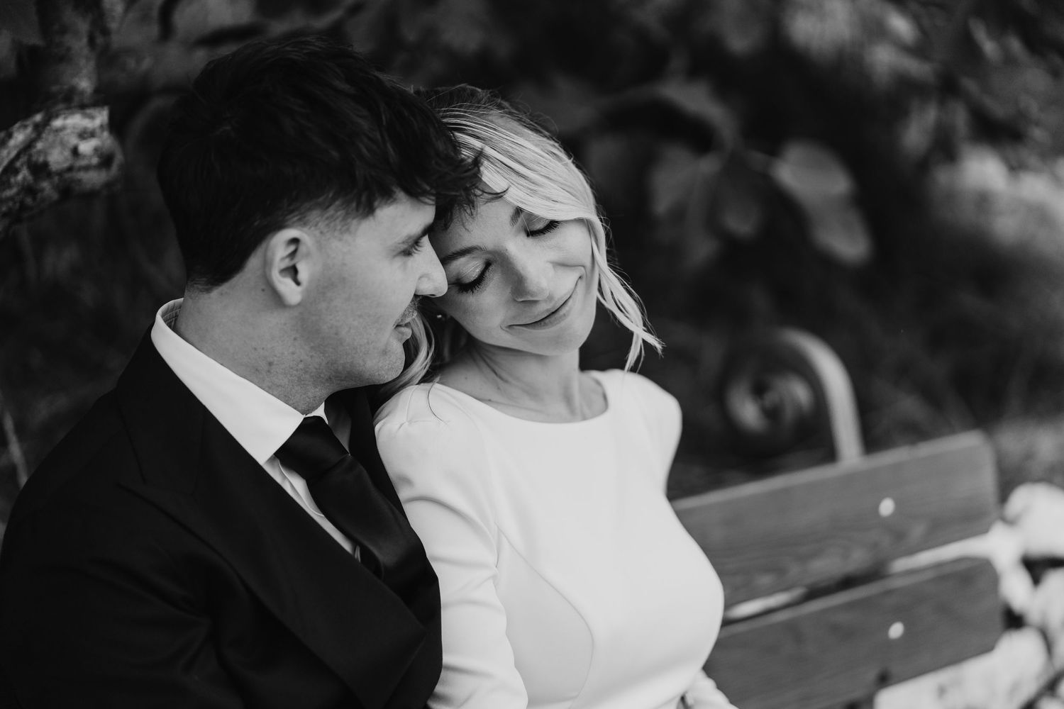 Black and white portrait of bride and groom sitting closely together on a bench in tender moment.
