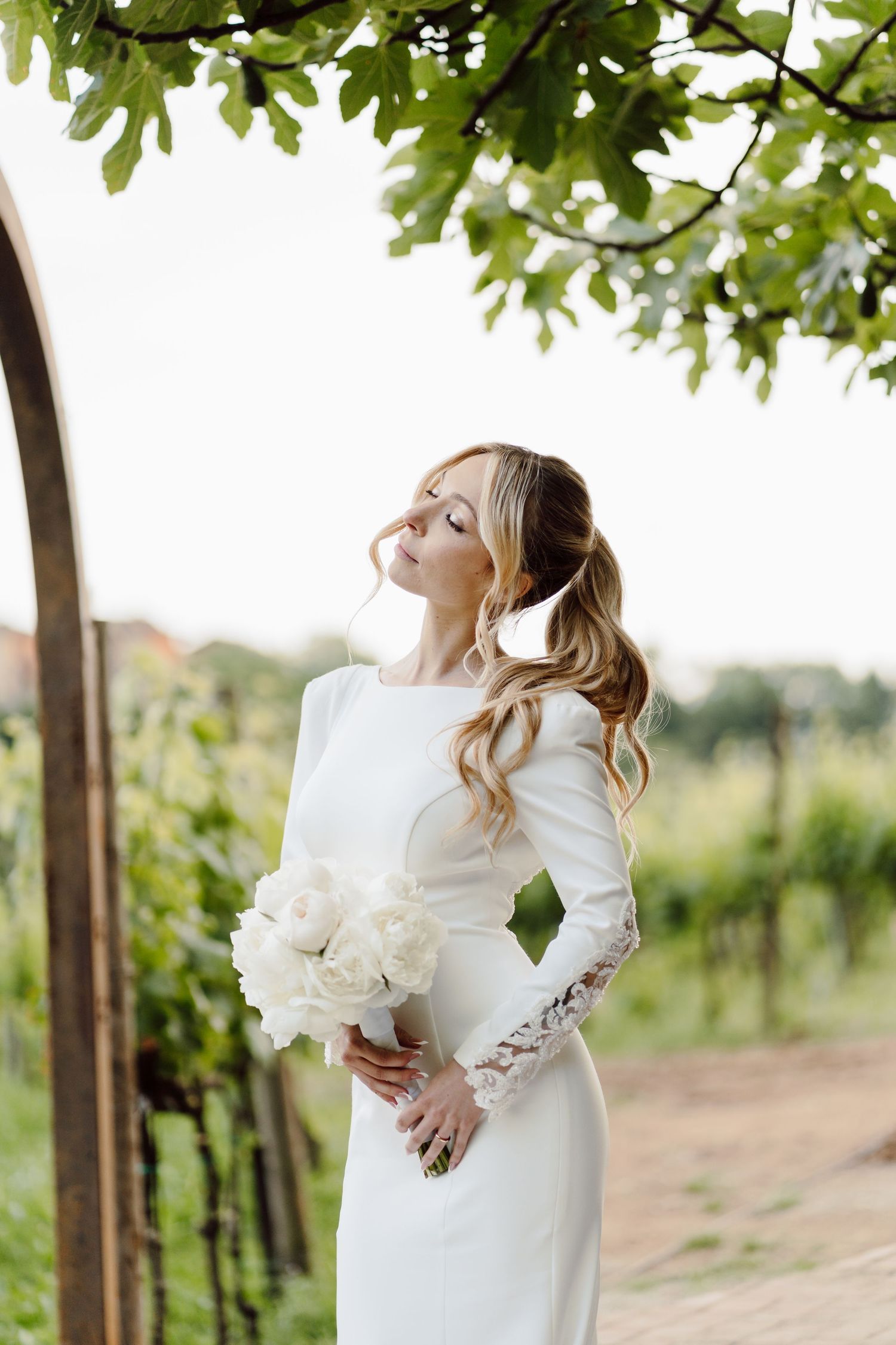 Elegant bride in long-sleeve white gown holding white peony bouquet beneath green leafy archway.