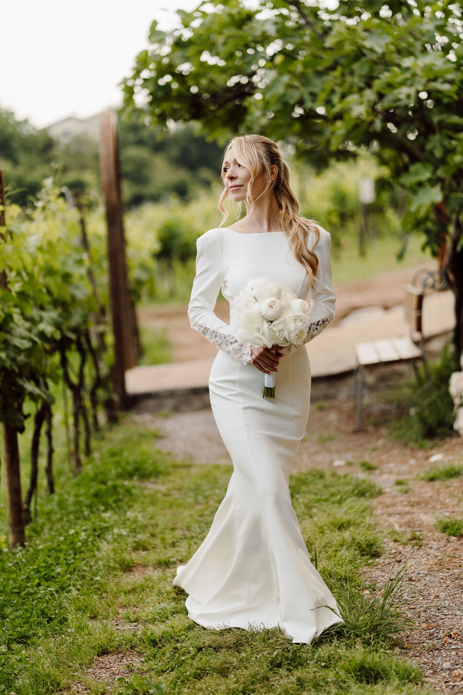 Bride in fitted long-sleeve white wedding dress carrying white bouquet walking through vineyard path.