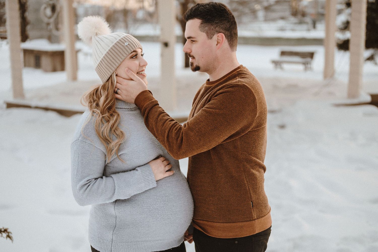 Intimate winter maternity photo of couple in warm sweaters standing in snowy setting.