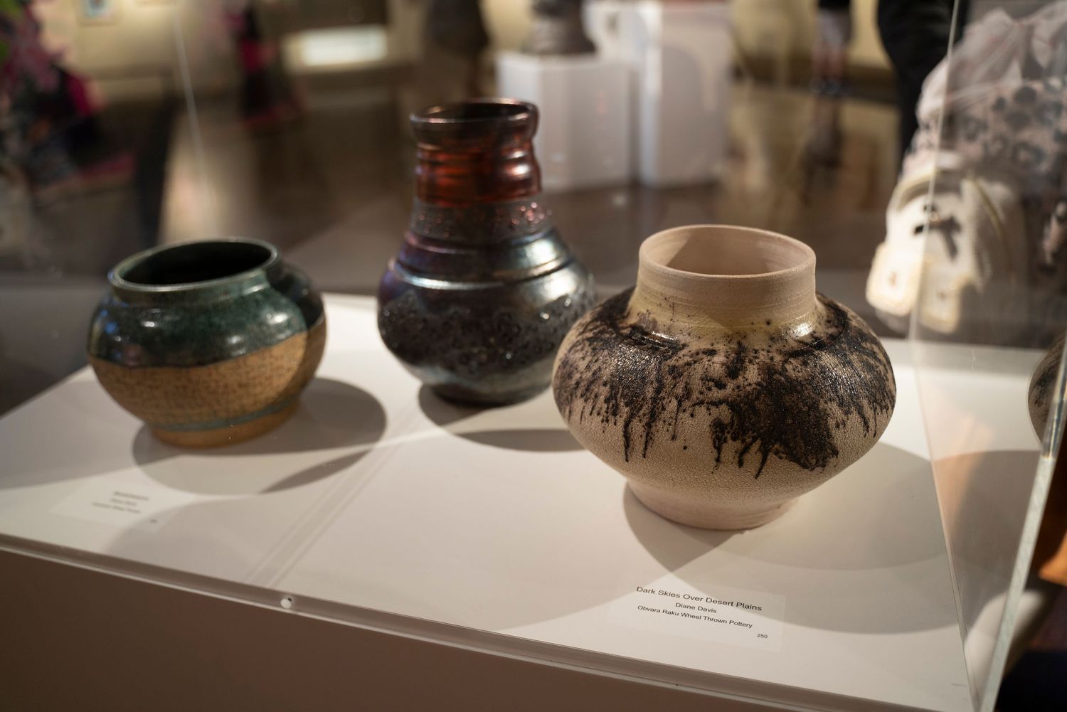 Display of three decorative ceramic pots with rustic designs and varied earth-tone glazes on white pedestal.