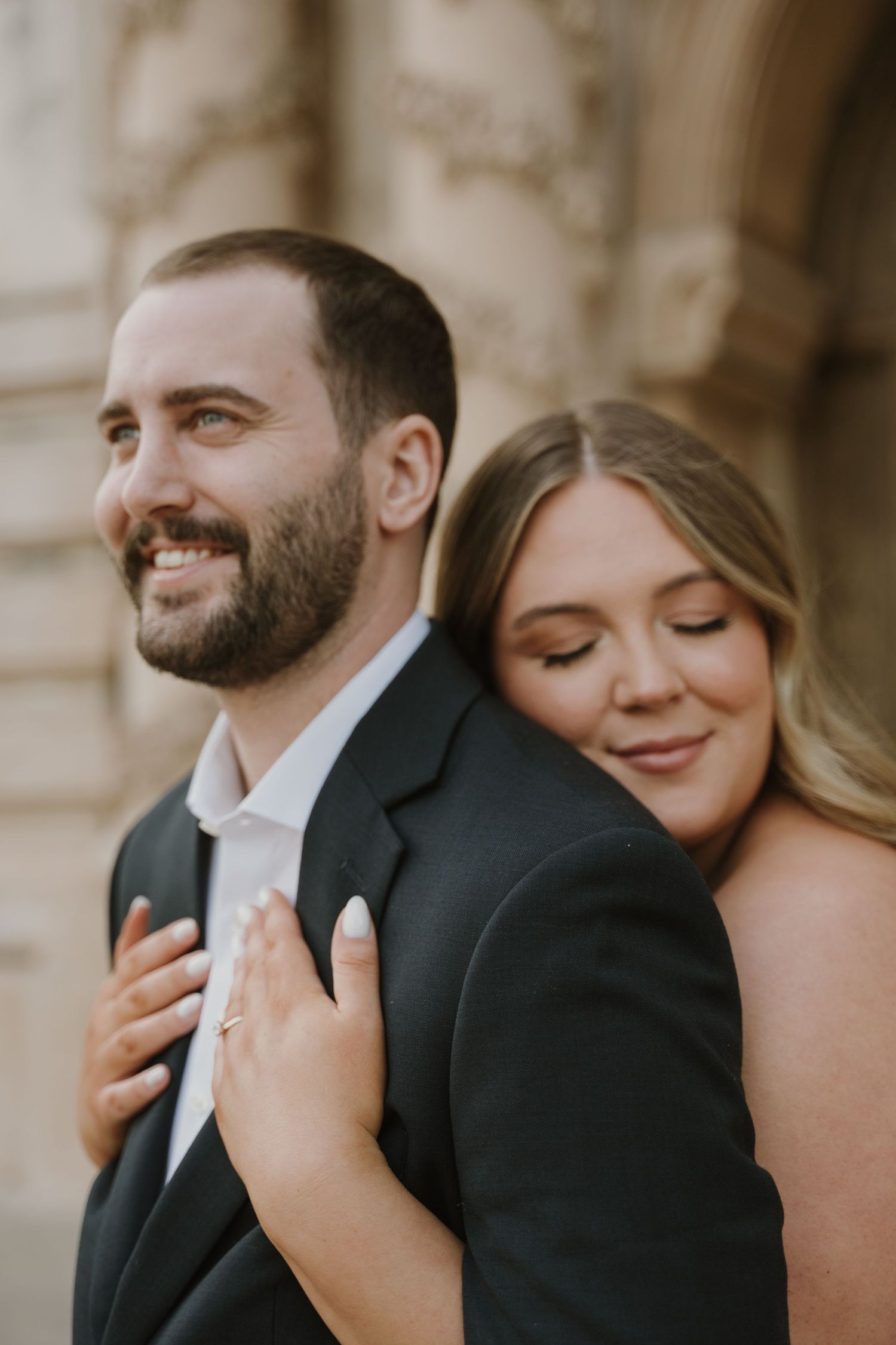 A romantic engagement photo of a couple embracing against a stone building exterior.