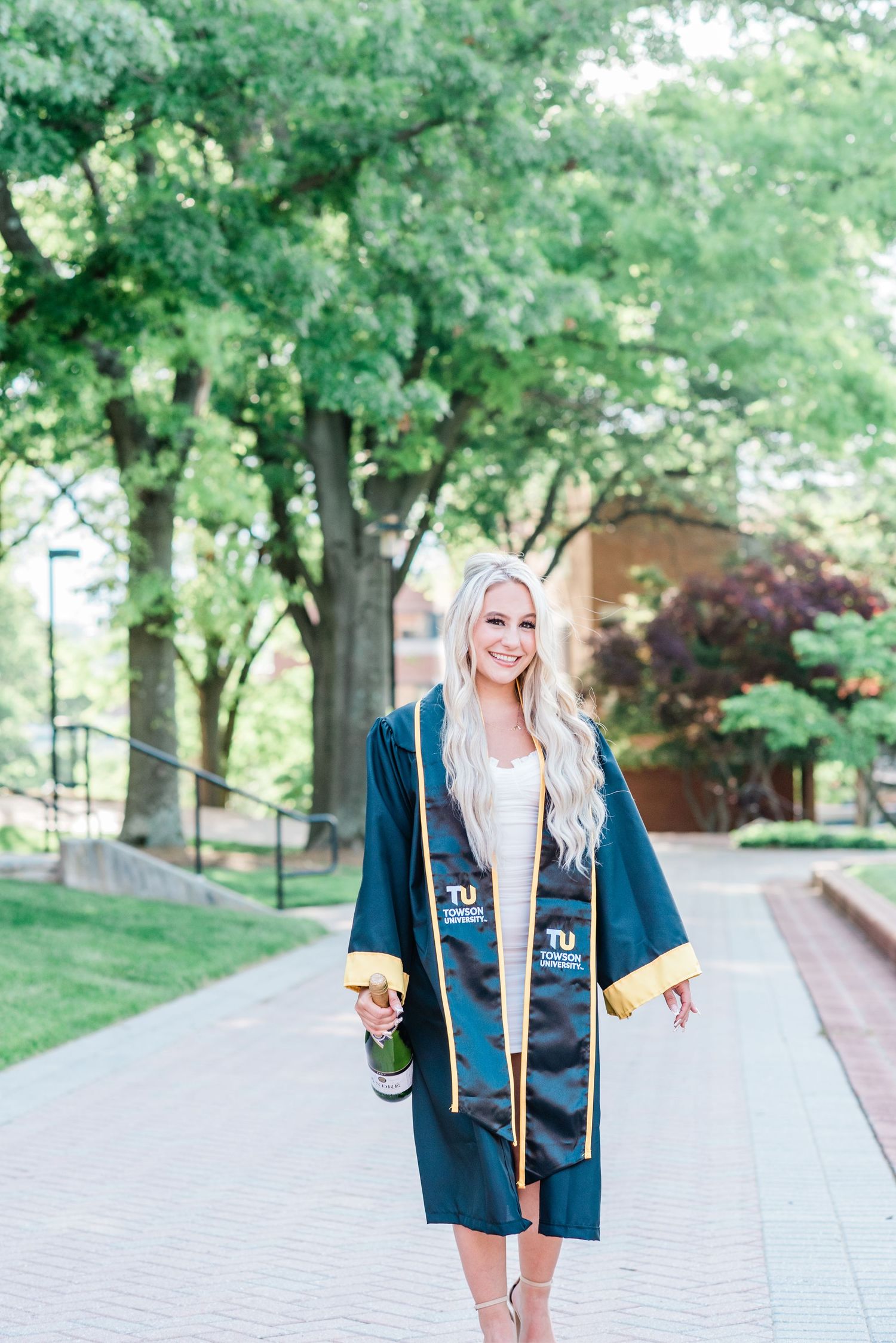 Graduate in cap and gown stands on campus walkway surrounded by green trees.