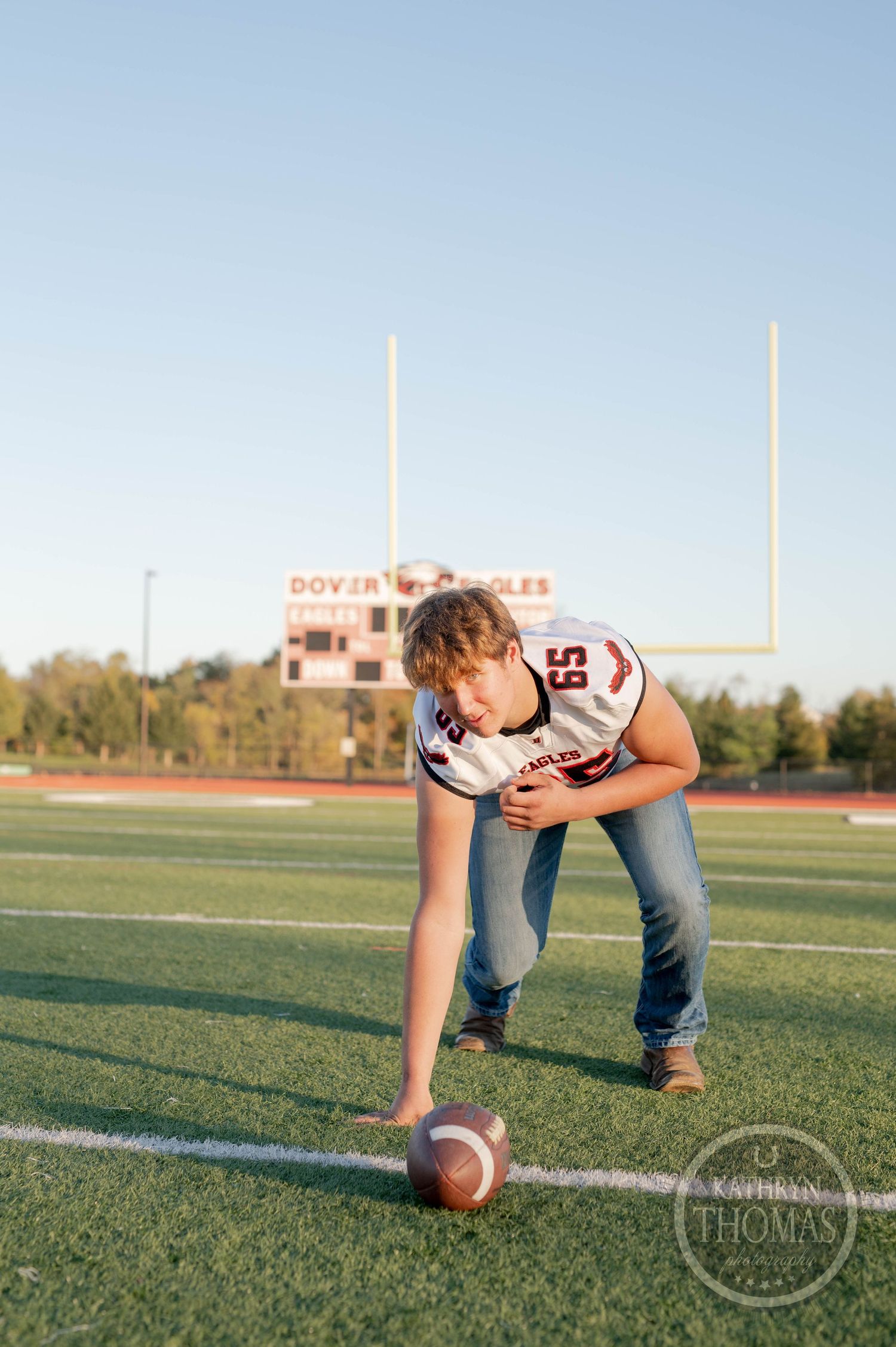 Football player in uniform demonstrates three-point stance with ball on field with goal posts visible.