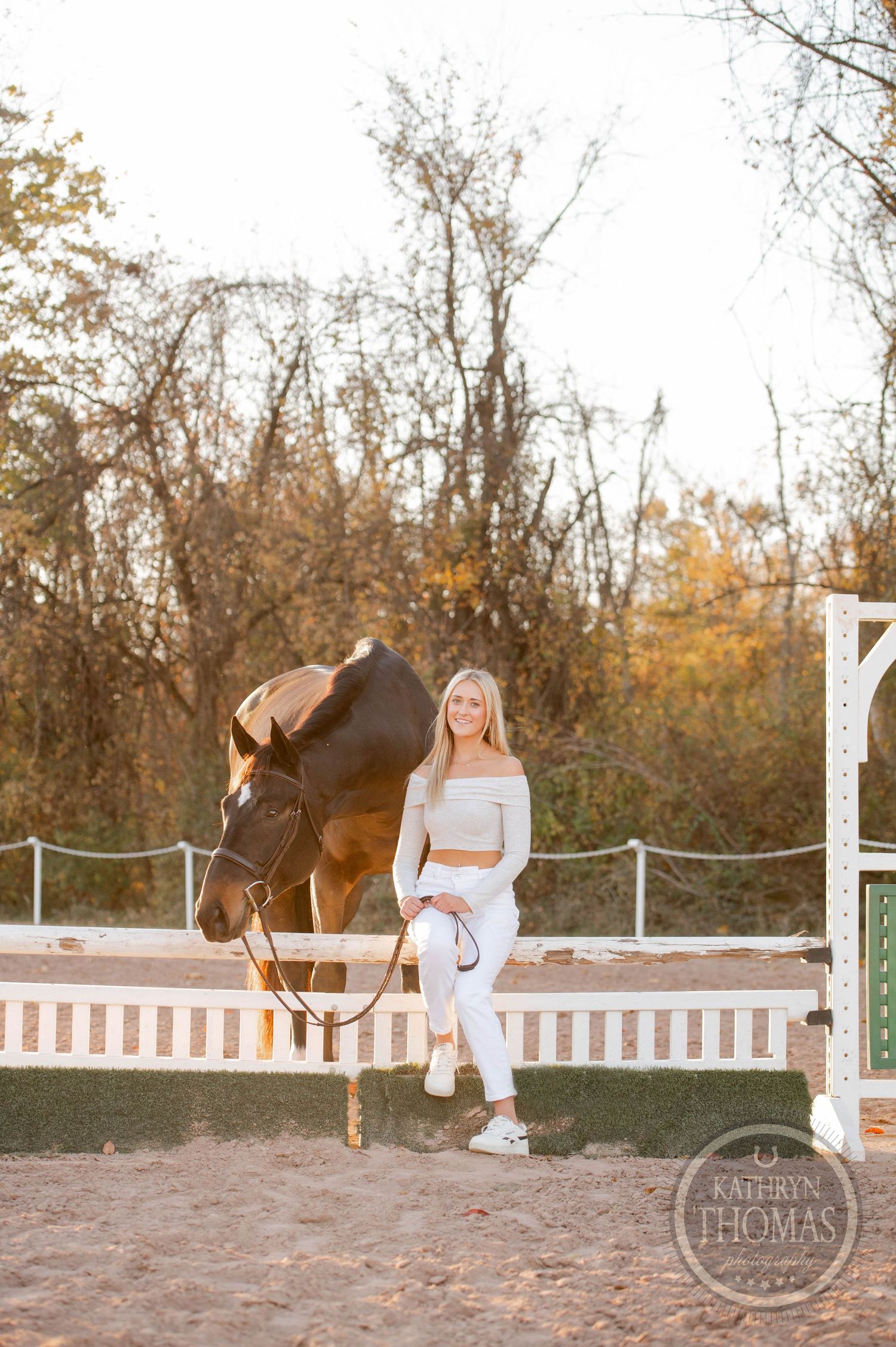 A bay horse jumps over a white fence post while someone in white stands nearby at sunset.