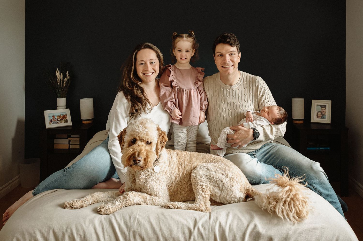 A cozy family portrait on a bed features a large goldendoodle dog alongside people in casual attire.