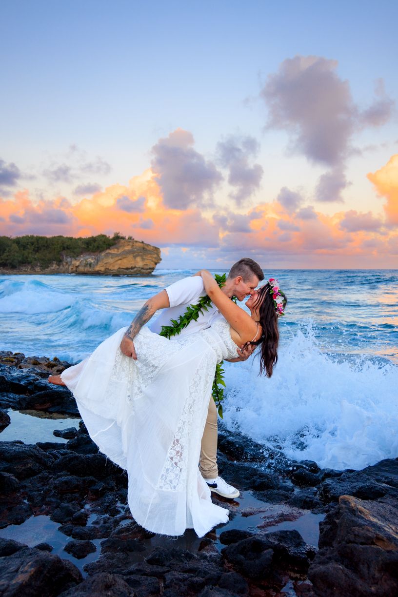 A romantic wedding couple dips on rocky Hawaiian coastline at sunset as waves crash behind them.