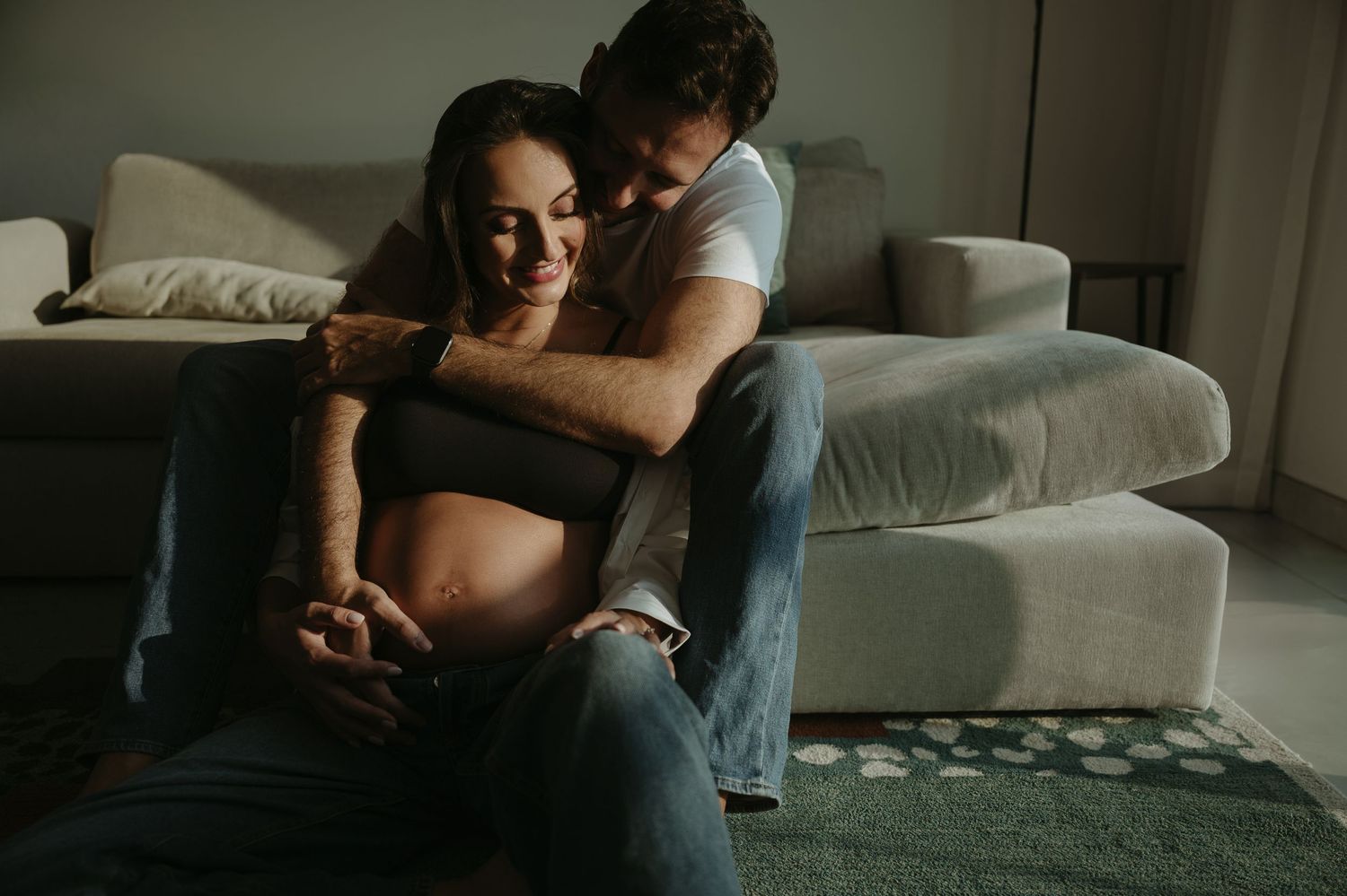 Expecting mother relaxes on geometric rug by window in casual home maternity photo.