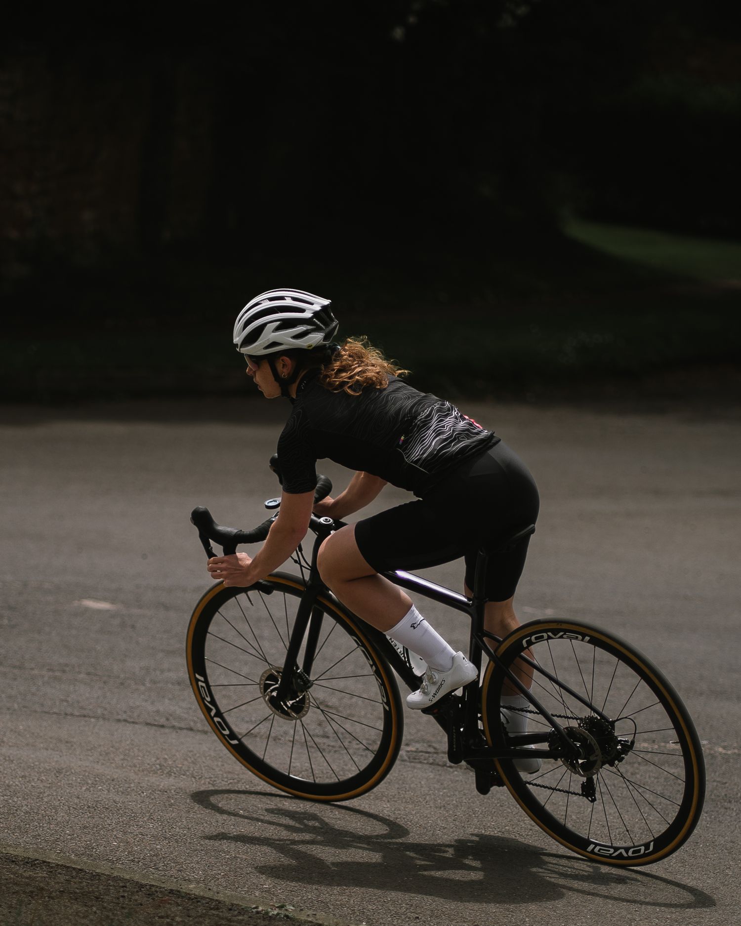Cyclist wearing black cycling gear and white helmet rides a road bike on dark pavement in moody lighting.