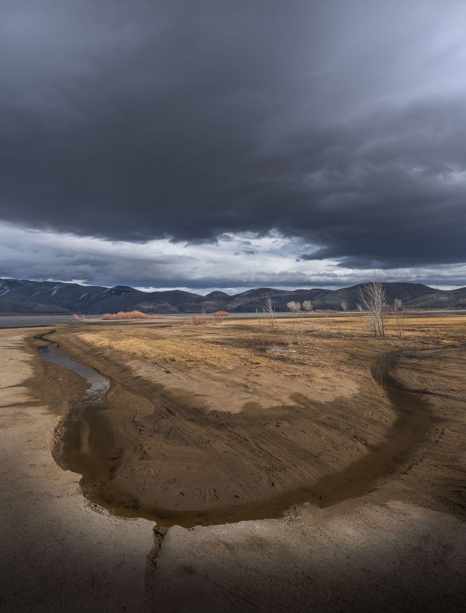 Dramatic dark storm clouds loom over a winding dry riverbed through arid terrain with mountains in the distance.