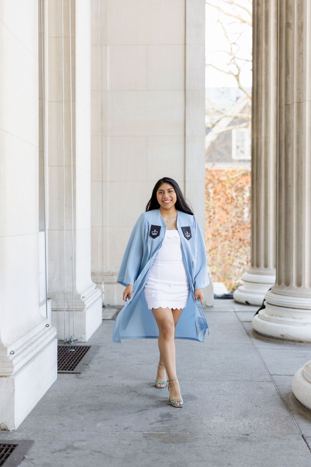 Graduate in light blue academic gown walking confidently through columned corridor.