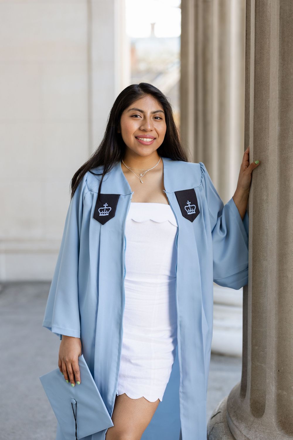 Graduate in light blue academic regalia posing between classical columns.