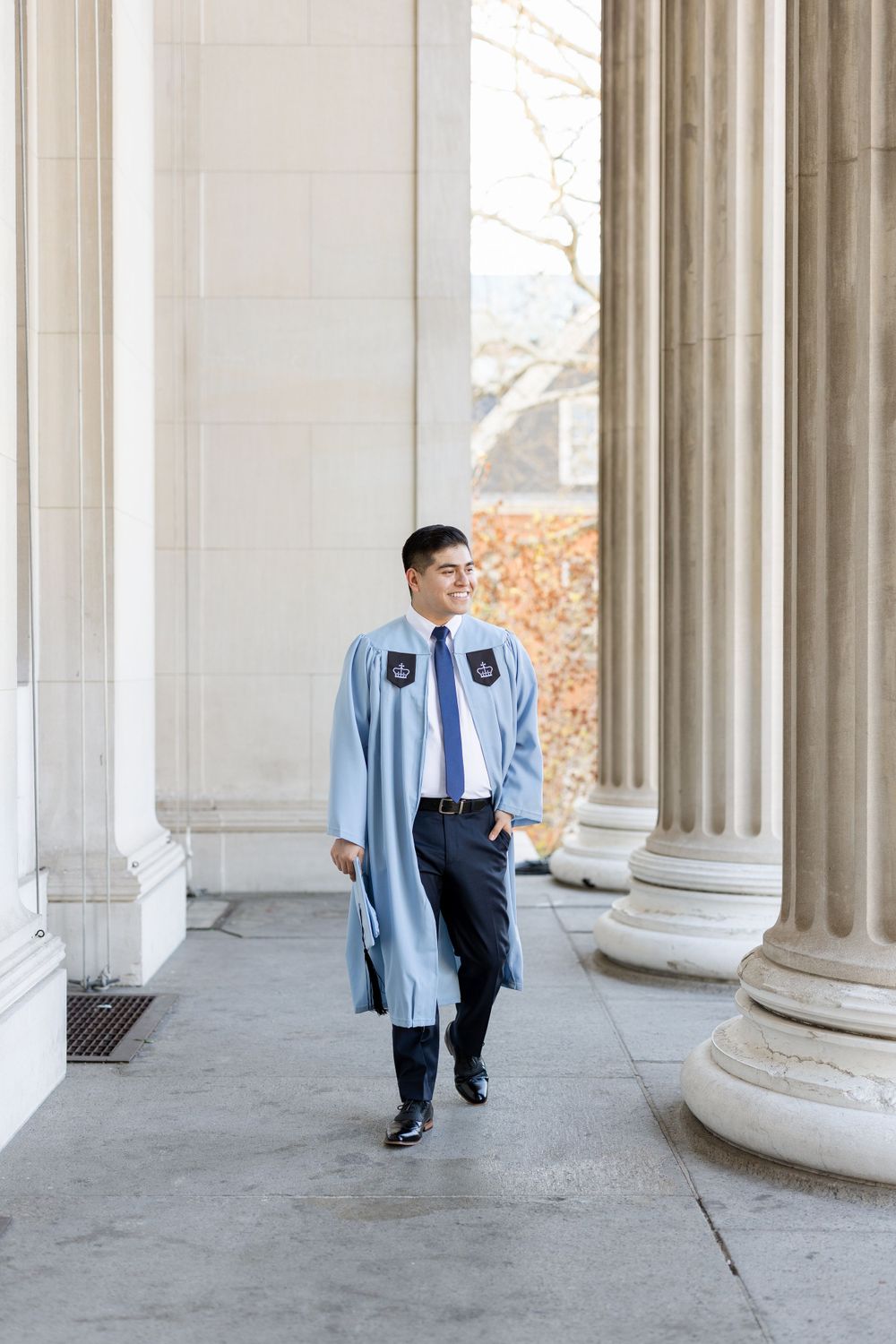 Student wearing light blue graduation gown walks between white stone pillars.