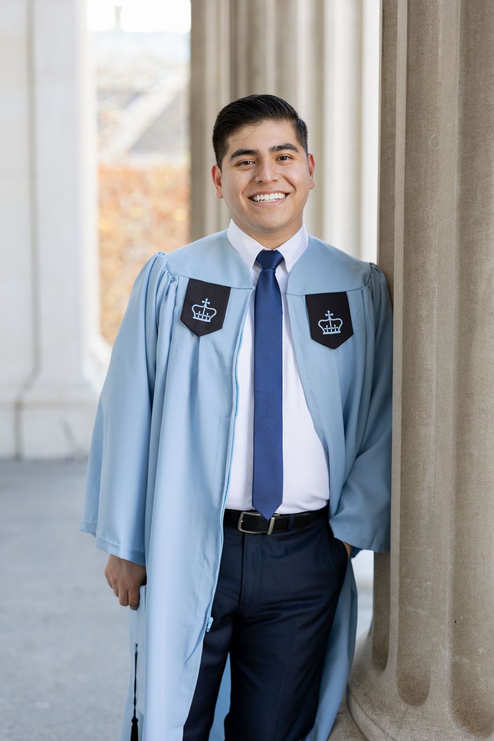 Graduate in Columbia University regalia with crown emblems stands by classical columns.