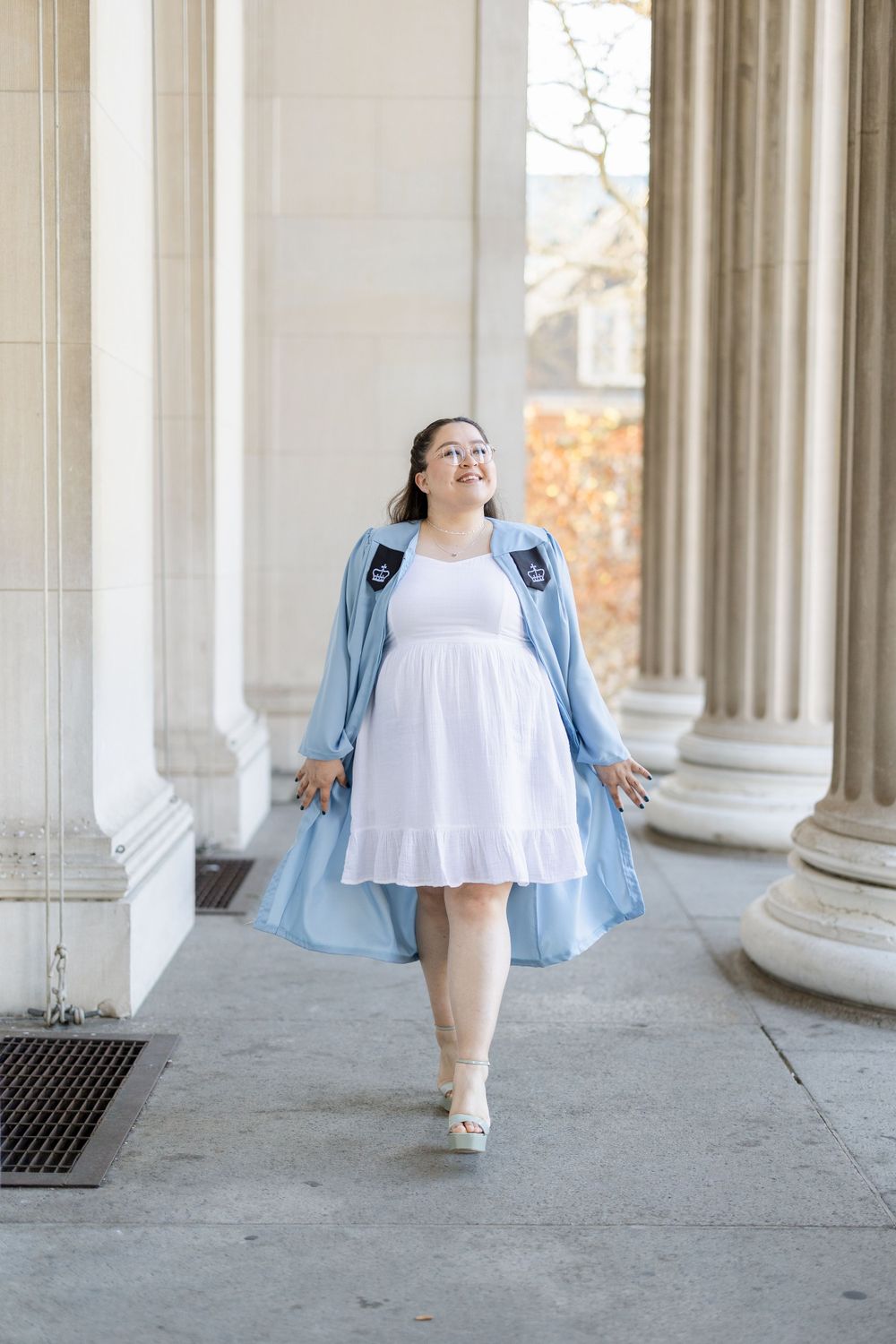 Graduate in white dress and light blue gown twirling between classical columns.