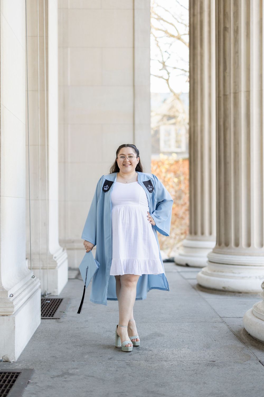 Graduate in light blue academic regalia poses between classical columns on campus.