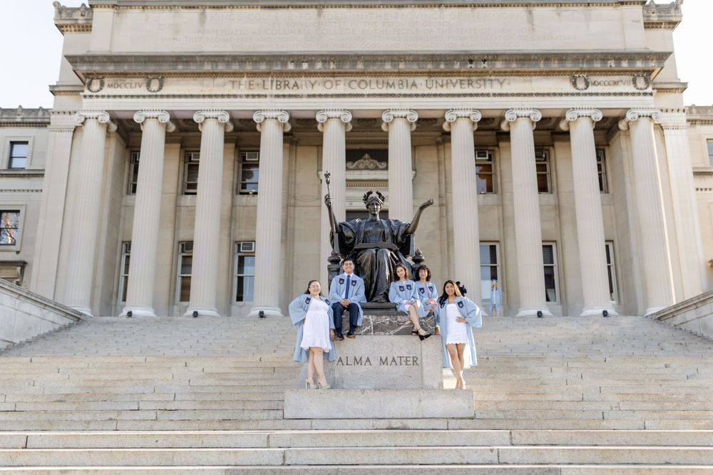 Graduates pose by the Alma Mater statue on the steps of a grand columned university building.