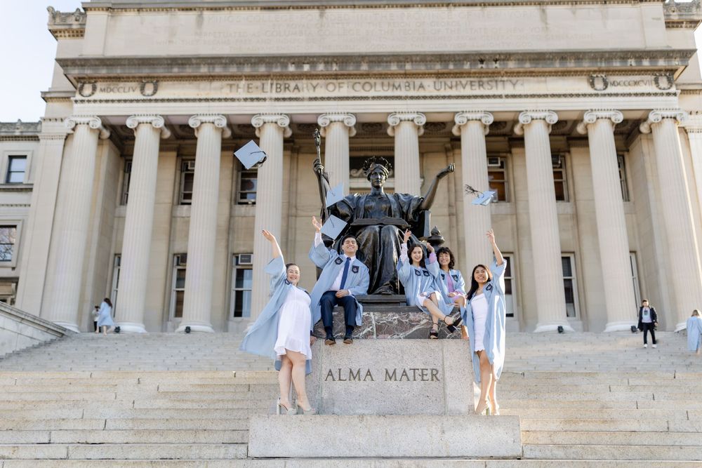 Students celebrate graduation with creative poses around the Alma Mater statue and neoclassical architecture.