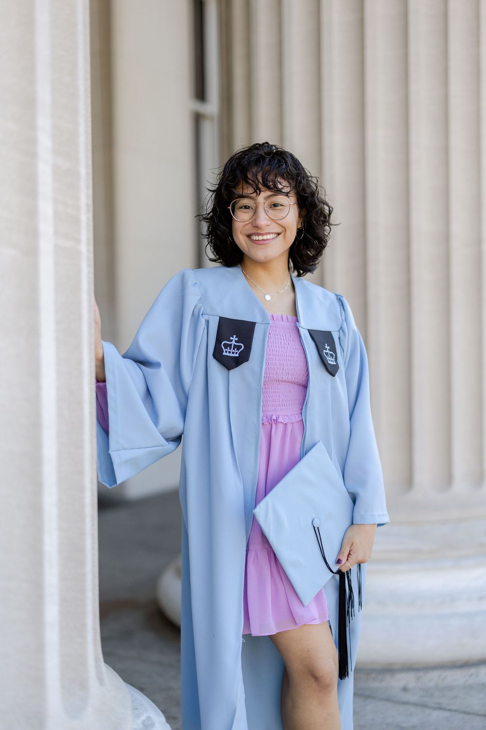 Graduate in light blue regalia and pink dress poses between classical columns.