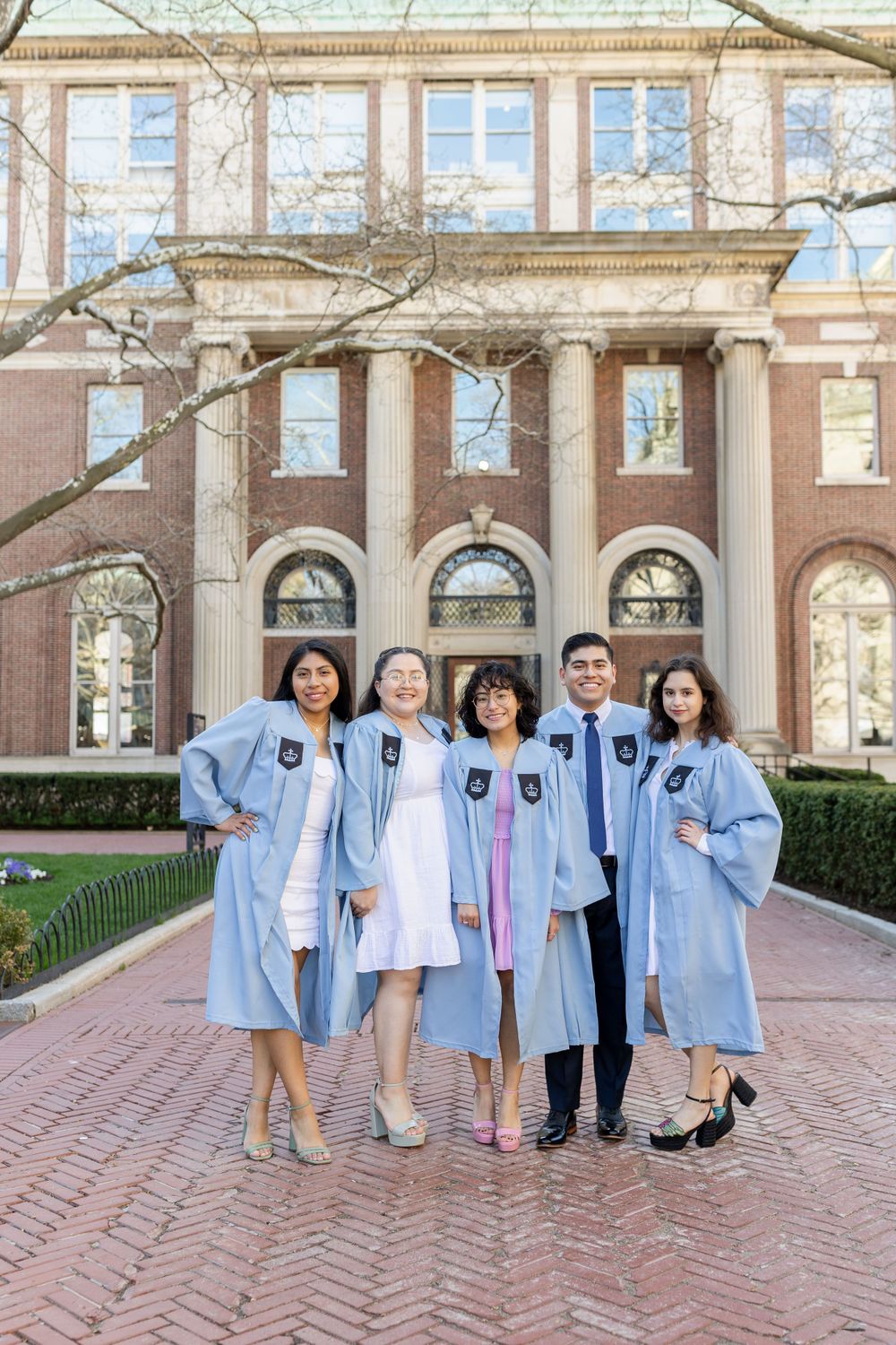 Graduates in light blue academic regalia pose in front of a brick collegiate building with columns.