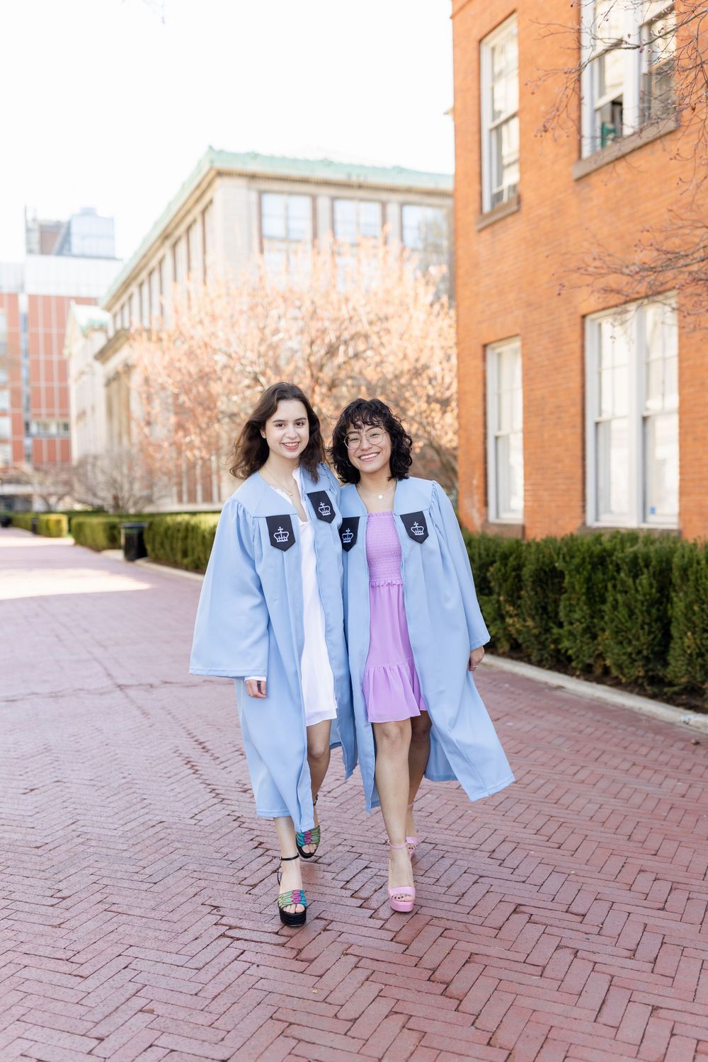 Two graduates in light blue academic robes walk along a brick pathway on campus.