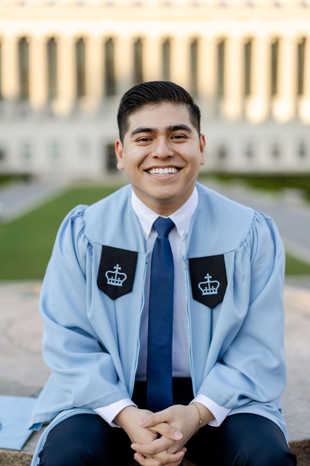 Columbia graduate in light blue regalia sitting outdoors on campus.