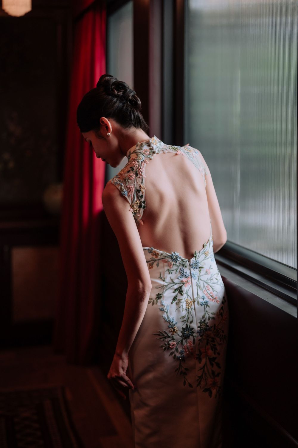 Elegant backless qipao dress with floral embroidery details shown from behind near a window with red curtains.