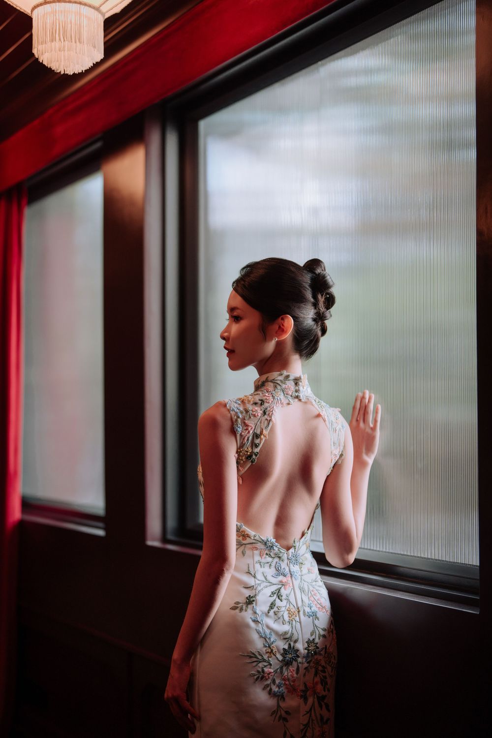 Elegant backless qipao dress with floral embroidery shown in moody lighting against frosted window with red accents.