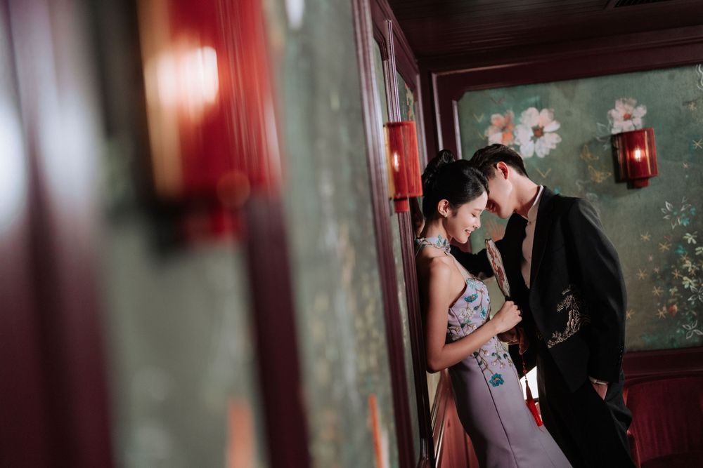 A romantic couple embraces in a dimly lit hallway with red walls and decorative floral wallpaper.