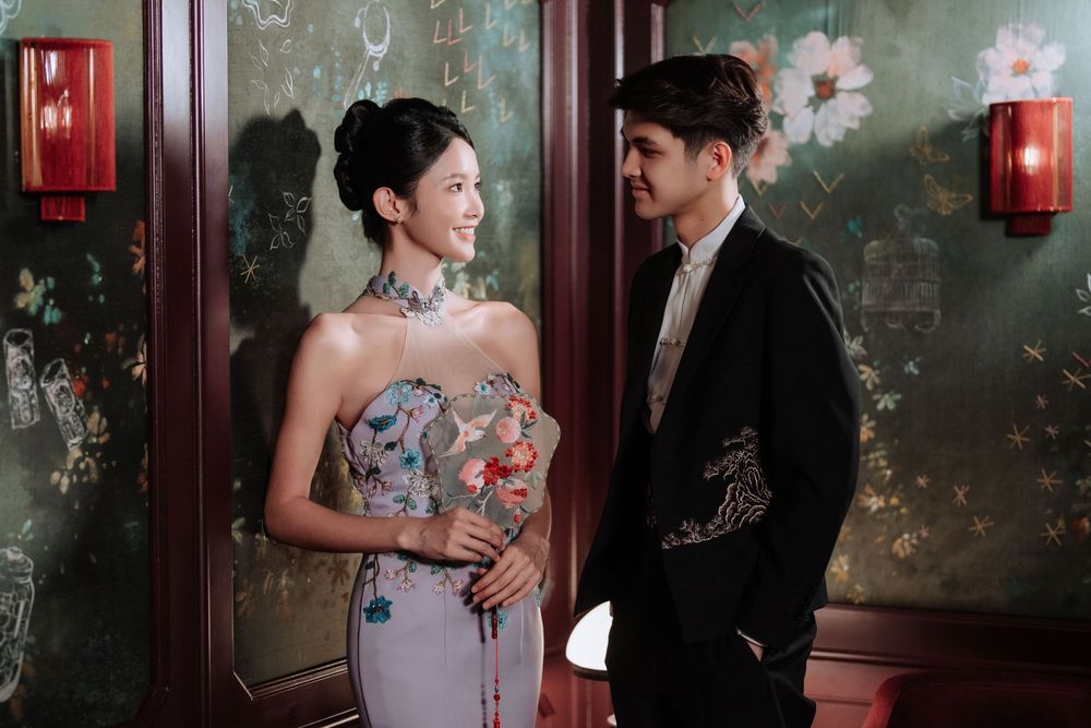 A couple in formal attire stands together against a textured wall with red lanterns and floral decorations.