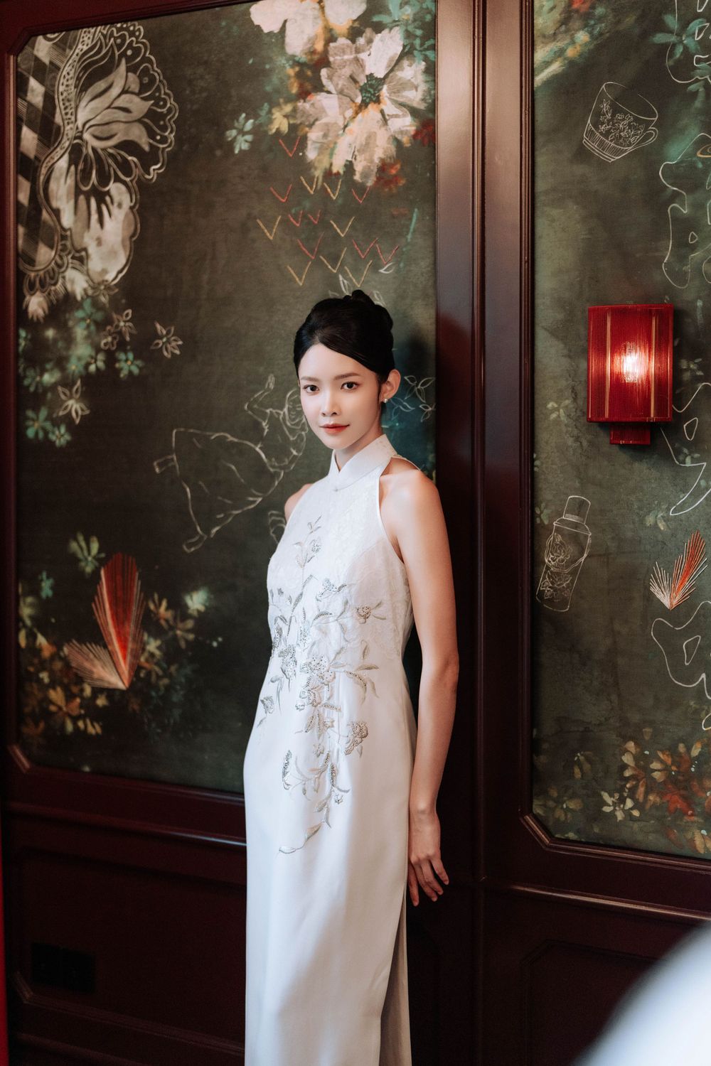 Elegant traditional Chinese cheongsam dress in white with floral embroidery against dark floral wallpaper and red lantern.