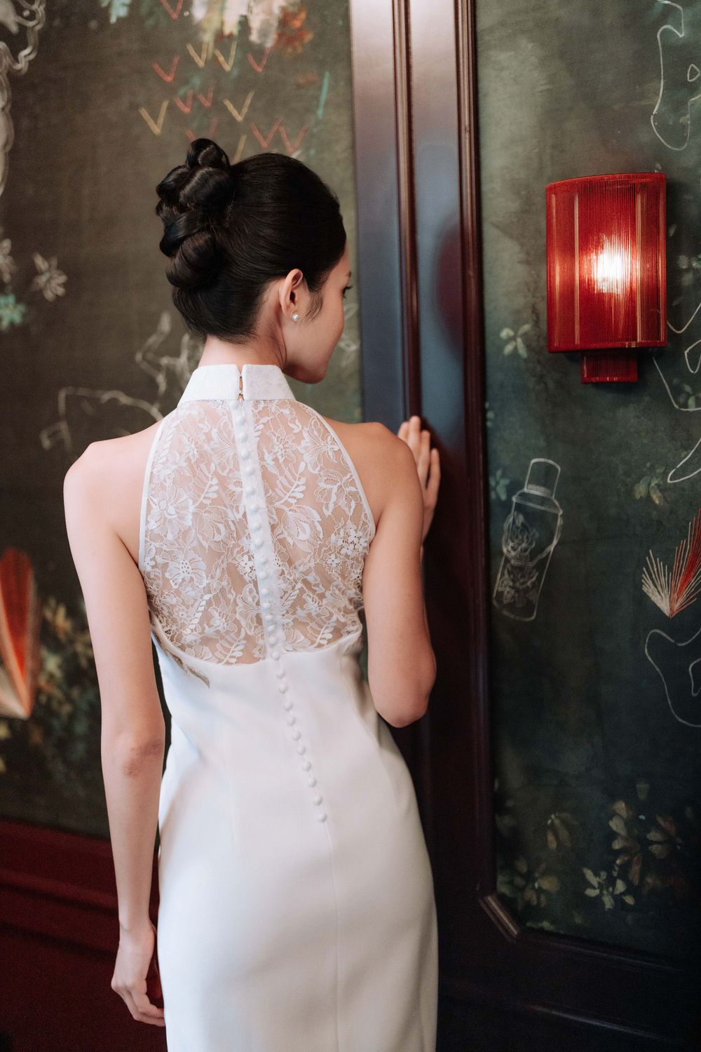 A white lace qipao dress with delicate sheer back details shown against a dark green wallpapered wall with red sconce.