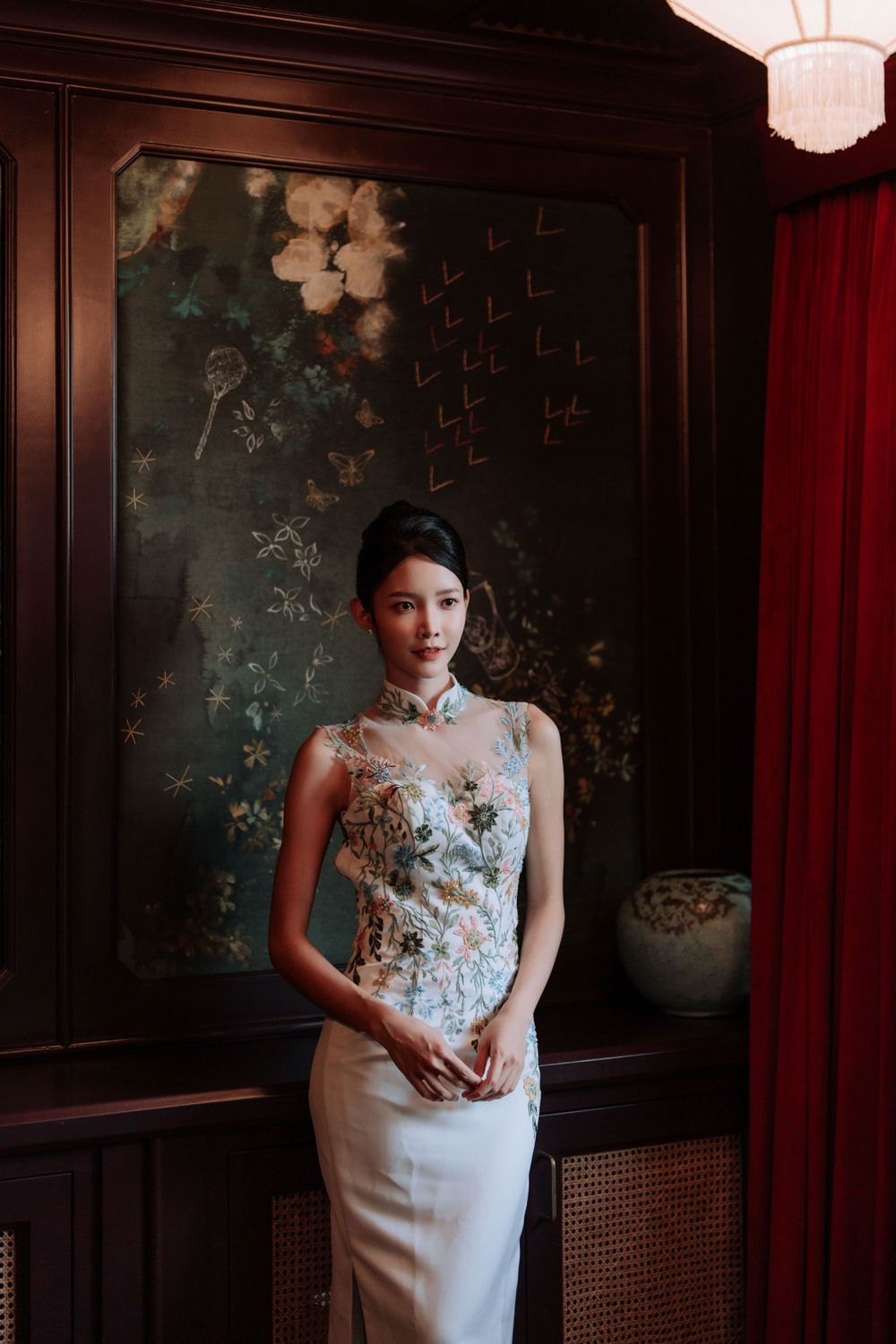 Elegant figure in a fitted white lace qipao dress poses against dark wood paneling and floral wallpaper.