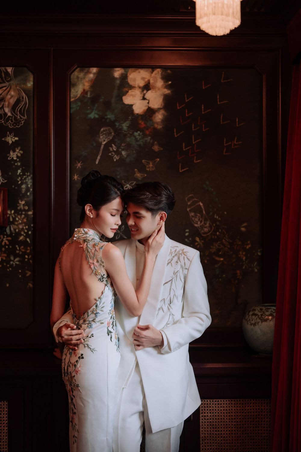 Intimate portrait of couple in traditional white Chinese wedding attire against dark floral backdrop.