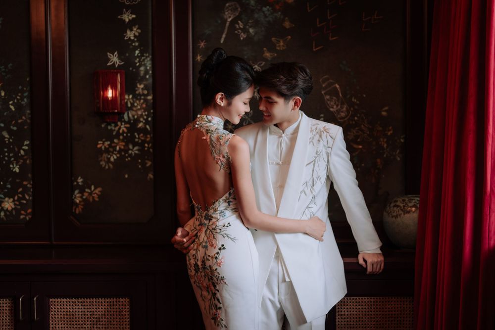 A romantic wedding couple in white attire embrace in an elegant room with antique wallpaper and red curtains.
