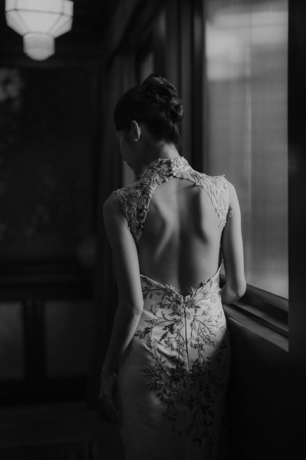 Black and white portrait of embroidered backless qipao dress shown in moody window lighting.