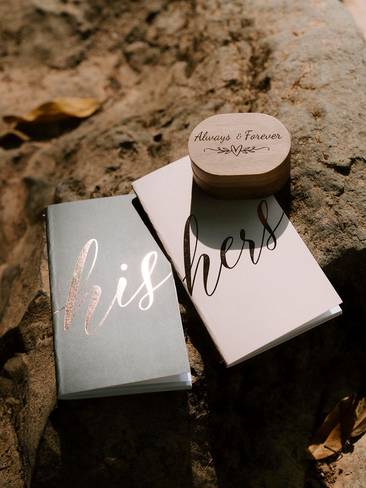 Wedding vow books with 'His' and 'Hers' text displayed on natural stone surface.